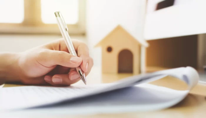 picture of a hand writing on a piece of paper with a wooden model of a house in the background