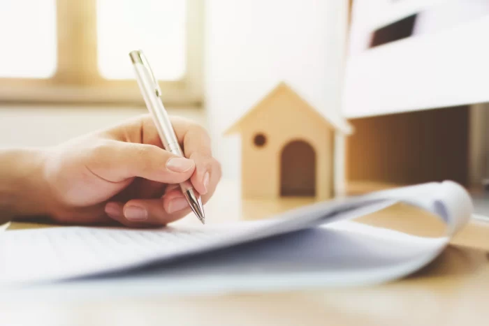 picture of a hand writing on a piece of paper with a wooden model of a house in the background