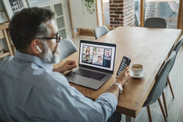 Image of man on in front of his laptop on a video call, whilst looking at his mobile phone.